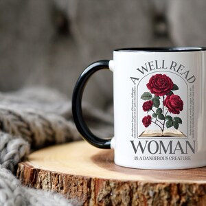 May include: White ceramic mug with a black handle and rim. The mug features a design of red roses and the text "A WELL READ WOMAN IS A DANGEROUS CREATURE". The mug is on a wooden surface.