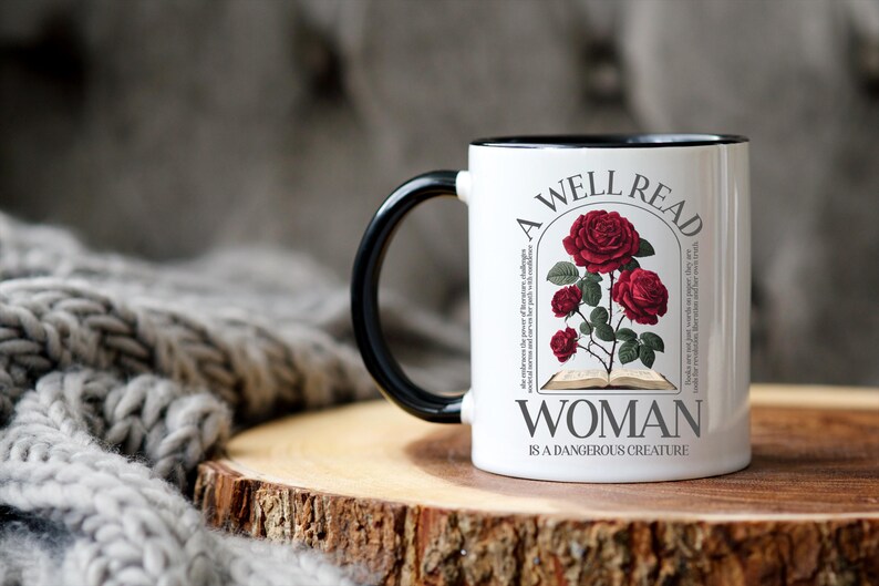 May include: White ceramic mug with a black handle and rim. The mug features a design of red roses and the text "A WELL READ WOMAN IS A DANGEROUS CREATURE". The mug is on a wooden surface.