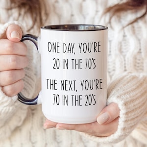 Puede incluir: Taza de cer&aacute;mica blanca con interior y asa negros, con el texto negro "ONE DAY, YOU'RE 20 IN THE 70'S THE NEXT, YOU'RE 70 IN THE 20'S". La taza la sostiene una persona que lleva un jersey color crema.