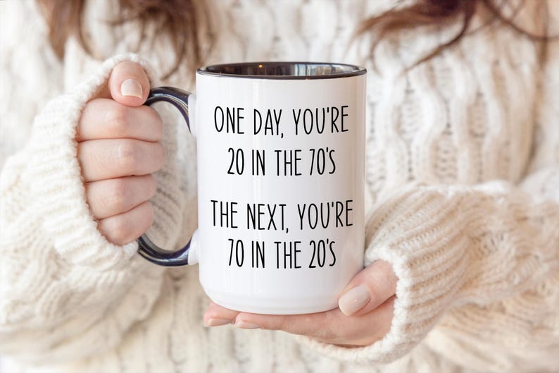 Puede incluir: Taza de cer&aacute;mica blanca con interior y asa negros, con el texto negro "ONE DAY, YOU'RE 20 IN THE 70'S THE NEXT, YOU'RE 70 IN THE 20'S". La taza la sostiene una persona que lleva un jersey color crema.