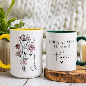 May include: Two white ceramic mugs. The left mug has a yellow handle and interior, featuring a black line-art floral bouquet with pink accents and the name Sarah. The right mug has a dark green handle and interior, with the text Look at you turning 25 twice and a small black heart.