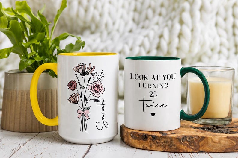 May include: Two white ceramic mugs. The left mug has a yellow handle and interior, featuring a black line-art floral bouquet with pink accents and the name Sarah. The right mug has a dark green handle and interior, with the text Look at you turning 25 twice and a small black heart.