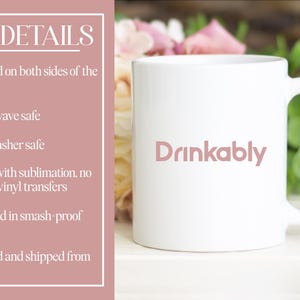 May include: White ceramic mug with the word "Drinkably" in a soft pink font. The mug has a curved handle and is set against a background of flowers and greenery. The mug is microwave and dishwasher safe.