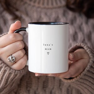 May include: White ceramic mug with black interior and handle, featuring the text "luna's mum" and a heart symbol. The mug is held by a person wearing a brown sweater.