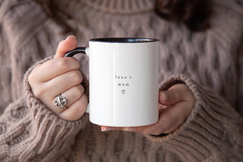 May include: White ceramic mug with black interior and handle, featuring the text "luna's mum" and a heart symbol. The mug is held by a person wearing a brown sweater.