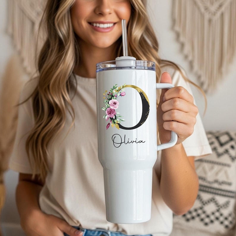 May include: White tumbler with a handle, a clear lid, and a silver straw. The tumbler features a black letter "O" with a gold accent and a floral design. The name "Olivia" is written in script below the letter.