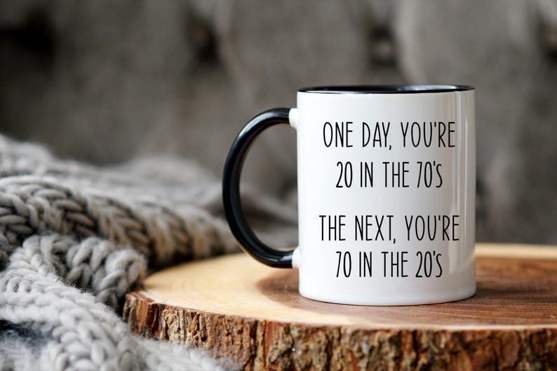 Puede incluir: Taza de cer&aacute;mica blanca con asa e interior negros. La taza presenta el texto "ONE DAY, YOU'RE 20 IN THE 70'S THE NEXT, YOU'RE 70 IN THE 20'S" en letras negras. La taza est&aacute; sobre una superficie de madera.