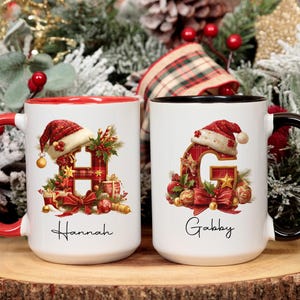 May include: Two white ceramic mugs with Christmas designs. One has a red handle and rim, the other a black handle and rim. Each mug features a letter decorated with festive elements, a Santa hat, and the names "Hannah" and "Gabby".