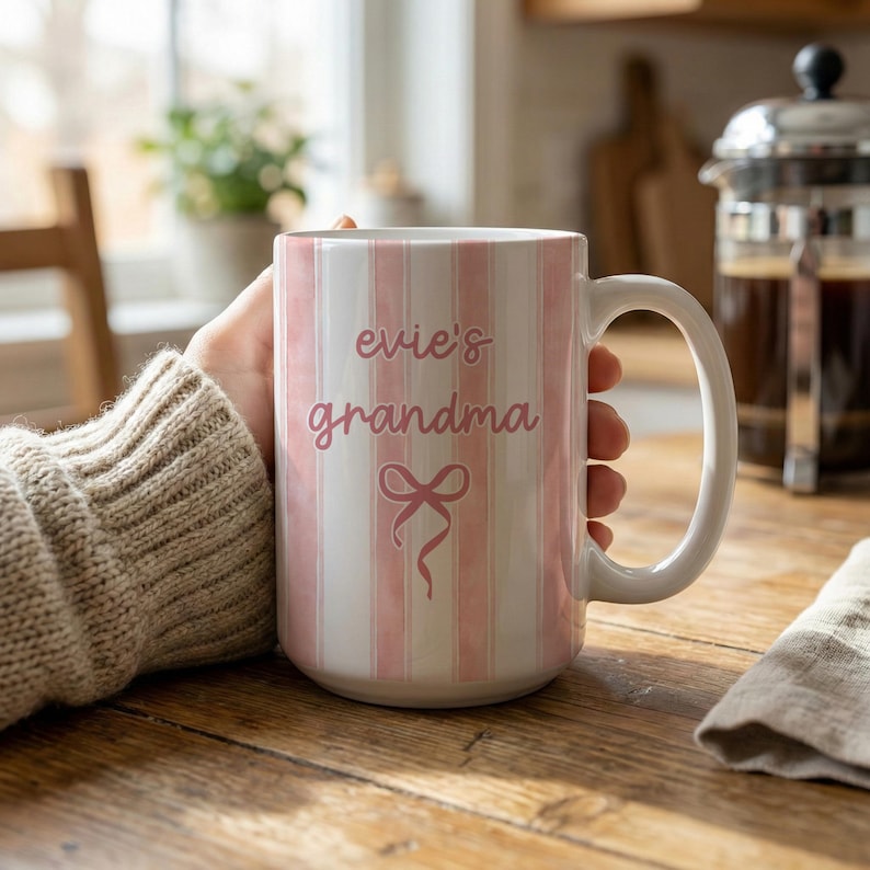 May include: A white and pink striped ceramic mug with the words "evie's grandma" in pink script, along with a pink bow graphic. The mug has a large handle and is held by a person.