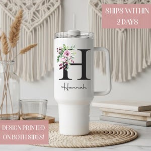 May include: White tumbler with a handle, floral design, and the letter "H". The floral arrangement includes pink and purple flowers with green leaves. The name "Hannah" is printed below the letter. The tumbler has a clear lid and a straw.