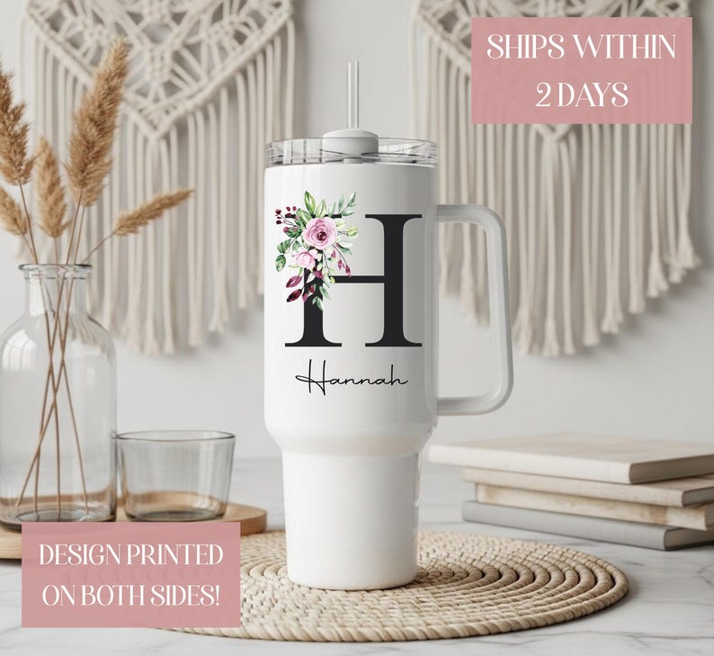 May include: White tumbler with a handle, floral design, and the letter "H". The floral arrangement includes pink and purple flowers with green leaves. The name "Hannah" is printed below the letter. The tumbler has a clear lid and a straw.