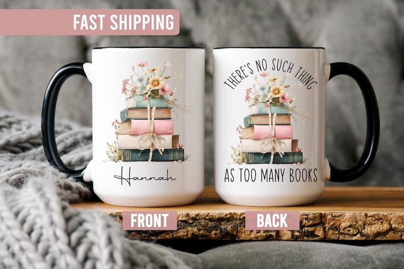 May include: White ceramic mug with a black handle and rim, featuring a stack of books adorned with flowers. The front of the mug has the name "Hannah" and the back says "THERE'S NO SUCH THING AS TOO MANY BOOKS."
