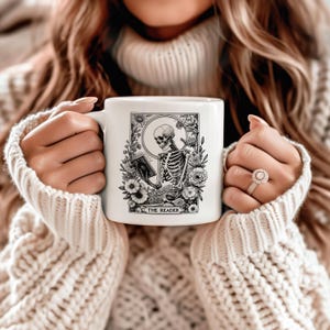 Puede incluir: Taza de cer&aacute;mica blanca con una ilustraci&oacute;n en blanco y negro de un esqueleto leyendo un libro, rodeado de flores. El texto "THE READER" est&aacute; en la parte inferior. La taza es sostenida por una persona que lleva un su&eacute;ter color crema.