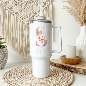 May include: White insulated tumbler with a handle and a straw. The tumbler features a rose gold letter "S" with a floral design and the name "Sophie" in script. The tumbler is designed for cold beverages.