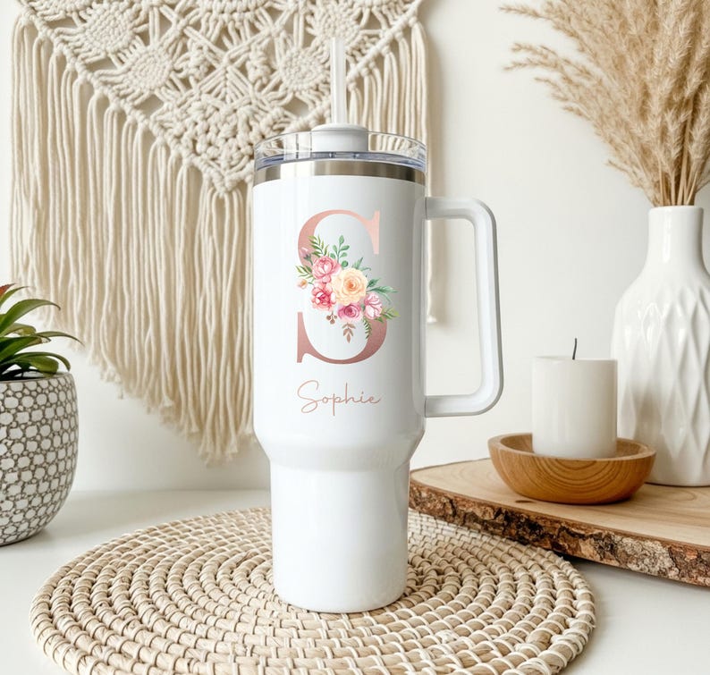 May include: White insulated tumbler with a handle and a straw. The tumbler features a rose gold letter "S" with a floral design and the name "Sophie" in script. The tumbler is designed for cold beverages.