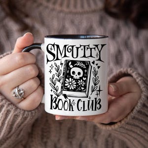 K&ouml;nnte beinhalten: Wei&szlig;er Keramikbecher mit schwarzem Henkel und Rand, mit dem Text "SMUTTY BOOK CLUB" in gotischer Schrift. Das Design des Bechers umfasst ein Buch mit einem Totenkopf und floralen Illustrationen.