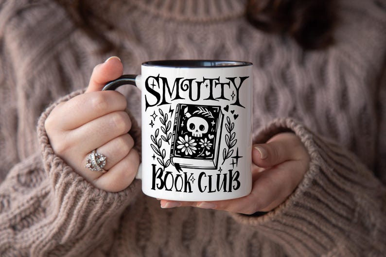 K&ouml;nnte beinhalten: Wei&szlig;er Keramikbecher mit schwarzem Henkel und Rand, mit dem Text "SMUTTY BOOK CLUB" in gotischer Schrift. Das Design des Bechers umfasst ein Buch mit einem Totenkopf und floralen Illustrationen.