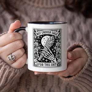 May include: White ceramic mug with a black handle and interior, featuring a black and white graphic design. The design includes a skeleton hand holding a bundle, a moon, stars, and the text "NOT ENOUGH SAGE FOR THIS SHIT."