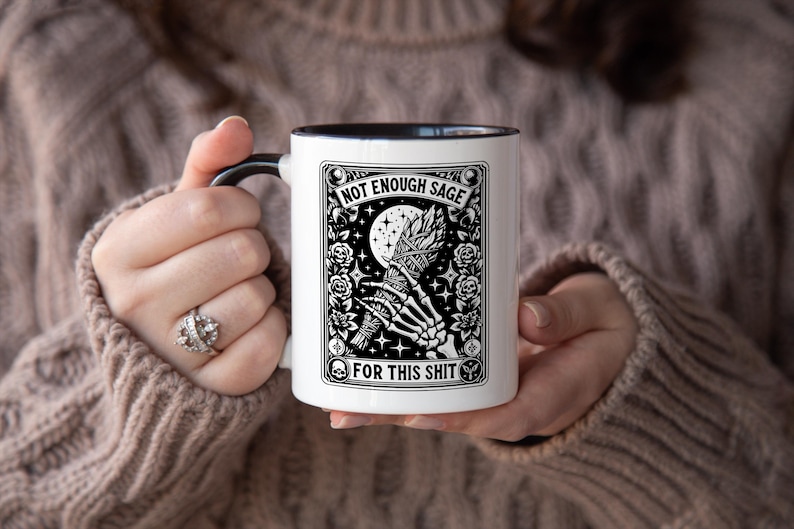 May include: White ceramic mug with a black handle and interior, featuring a black and white graphic design. The design includes a skeleton hand holding a bundle, a moon, stars, and the text "NOT ENOUGH SAGE FOR THIS SHIT."