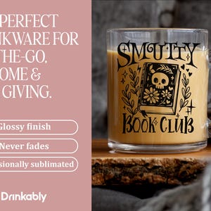 K&ouml;nnte beinhalten: Ein klarer Glasbecher mit Henkel, gef&uuml;llt mit einer hellbraunen Fl&uuml;ssigkeit. Der Becher hat schwarzen Text und eine Grafik eines Buches mit einem Totenkopf auf dem Cover, mit den Worten "SMUTTY BOOK CLUB".