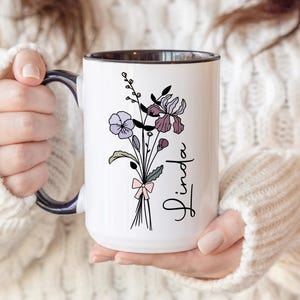 May include: White ceramic mug with a black interior and handle, featuring a floral bouquet design in purple, lavender, and burgundy hues. The name "Linda" is written in a script font. The mug is held by a person wearing a white sweater.