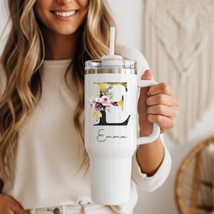 May include: White tumbler with a handle and a straw. The tumbler features a gold and black letter "E" design with floral accents and the name "Emma" in script. The tumbler is held by a person.