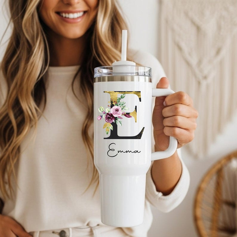 May include: White tumbler with a handle and a straw. The tumbler features a gold and black letter "E" design with floral accents and the name "Emma" in script. The tumbler is held by a person.
