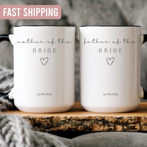 May include: Two white ceramic mugs with black handles and rims. One mug reads "mother of the BRIDE" and the other reads "father of the BRIDE", both with a heart symbol and the date 15.08.2025.