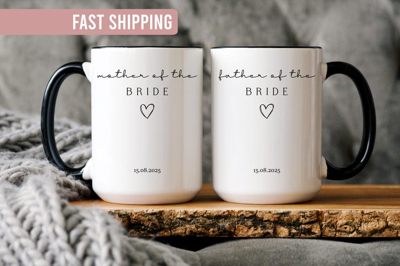 May include: Two white ceramic mugs with black handles and rims. One mug reads "mother of the BRIDE" and the other reads "father of the BRIDE", both with a heart symbol and the date 15.08.2025.