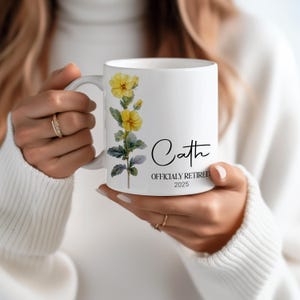 May include: White ceramic mug featuring a floral design with yellow flowers and green leaves. The mug has the name "Cath" in a script font, along with "OFFICIALLY RETIRED 2025". A perfect gift for a retiree.
