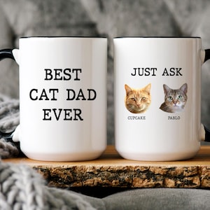May include: Two white ceramic mugs with black handles. The first mug features the black text BEST CAT DAD EVER. The second mug displays the black text JUST ASK above two cat portraits labelled CUPCAKE and PABLO in black font below each cat face.