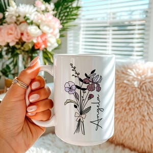 May include: A white ceramic mug featuring a black line art illustration of a wildflower bouquet tied with a small bow. The name Angela is printed in a black cursive script next to the flowers, which include purple and lavender accents.