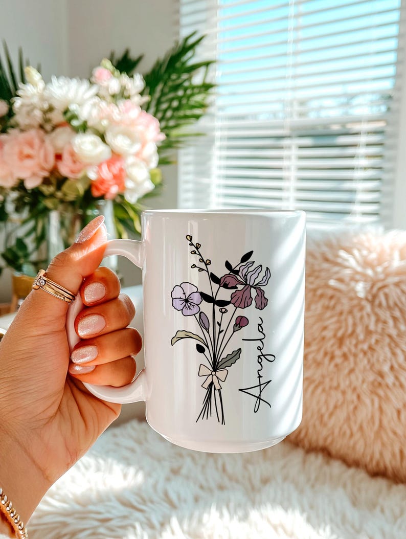 May include: A white ceramic mug featuring a black line art illustration of a wildflower bouquet tied with a small bow. The name Angela is printed in a black cursive script next to the flowers, which include purple and lavender accents.
