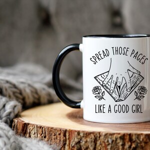 May include: White ceramic mug with a black handle and rim. The mug features the text "SPREAD THOSE PAGES LIKE A GOOD GIRL" with a graphic of hands holding a book and rose illustrations. The mug is on a wooden surface.