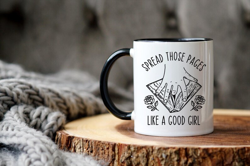 May include: White ceramic mug with a black handle and rim. The mug features the text "SPREAD THOSE PAGES LIKE A GOOD GIRL" with a graphic of hands holding a book and rose illustrations. The mug is on a wooden surface.