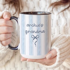 May include: A white ceramic mug with a black interior and handle, featuring blue vertical stripes and the text "archie's grandma" in a script font. A bow graphic is below the text. The mug is held by a person wearing a white sweater.