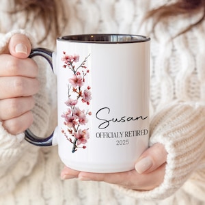 May include: White ceramic mug with a black interior and handle, featuring a floral design of pink blossoms. The mug is personalized with the name "Susan" and the text "OFFICIALLY RETIRED 2025".
