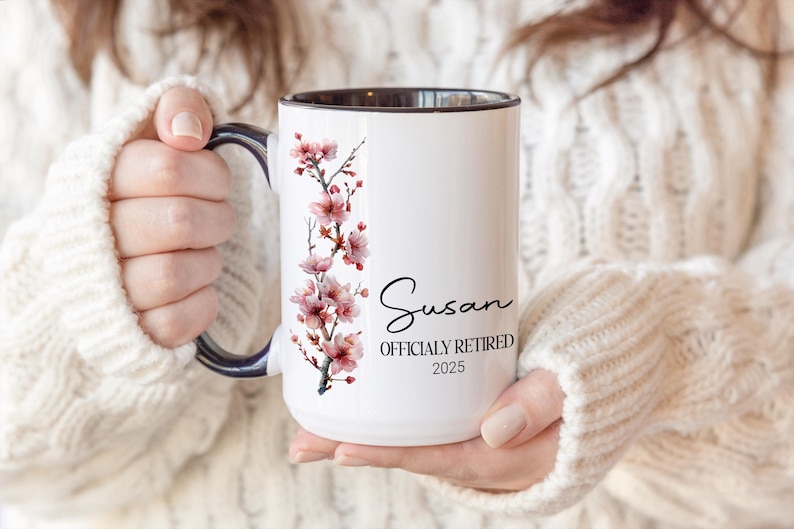 May include: White ceramic mug with a black interior and handle, featuring a floral design of pink blossoms. The mug is personalized with the name "Susan" and the text "OFFICIALLY RETIRED 2025".
