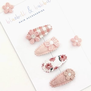 May include: A selection of pink and white hair clips and flower embellishments. The clips showcase floral and bow motifs, with a gingham design on one. The text "bluebell & bubba HAIR ACCESSORIES" is displayed on the card.