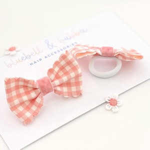 May include: A pink and white gingham hair bow with scalloped edges and a solid pink centre band, paired with a matching gingham hair tie on a white display card labelled bluebell & bubba HAIR ACCESSORIES with a small white flower icon.