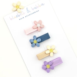 May include: Four hair clips on a white display card labelled bluebell & bubba HAIR ACCESSORIES. Each clip features a ribbed fabric base and an embroidered flower applique in pink, blue, beige, or purple with a contrasting centre.