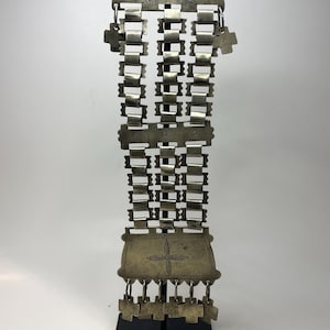 May include: A silver-toned metal decorative piece with an intricate design. The top features two birds, and the body has a series of rectangular and cross-shaped elements. The bottom has a rectangular piece with a floral design and dangling cross-shaped charms.