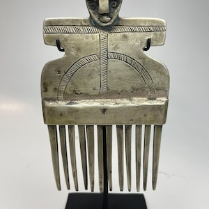 Puede incluir: Un peine de color plateado con un diseño estilizado de figura humana. El peine presenta una cara con una expresión sonriente y patrones geométricos. El peine está montado sobre un soporte negro.