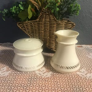 Puede incluir: Un azucarero color crema con tapa y una jarra de crema con boquilla. Ambas piezas tienen una banda plateada texturizada alrededor de la base. Una cesta tejida en forma de estrella con vegetación en el fondo.