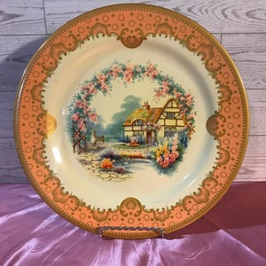 May include: A decorative plate with a cottage scene. The plate features a central image of a cottage surrounded by a floral arch. The rim is peach with gold accents. The plate is displayed on a metal stand.