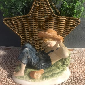 May include: A ceramic figurine of a child in overalls and a straw hat, resting on a patch of green grass. The figurine is on a white base, with a small loaf of bread nearby. A star-shaped woven basket with greenery is in the background.