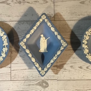 May include: Three blue and white decorative ceramic pieces. Two round plates and a diamond-shaped dish feature white relief designs and floral borders. The plates depict figures and a building. The diamond dish has a central statue.