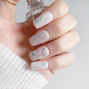 Puede incluir: Primer plano de una mano con uñas blancas de forma cuadrada adornadas con purpurina pastel y detalles iridiscentes. Las uñas tienen un acabado brillante. Se sostiene en la mano una botella de cristal transparente con tapón plateado. El texto "Chloemomo" es visible.