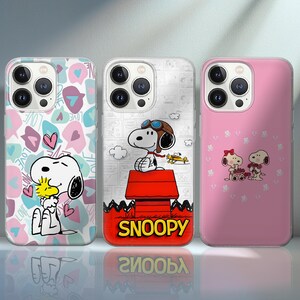 Pode incluir: Três capas de telemóvel com designs de Snoopy e Woodstock. Uma capa tem um padrão rosa e branco com Snoopy e Woodstock. Outra capa tem Snoopy com um chapéu de piloto numa casota vermelha com a palavra "SNOOPY". A terceira capa é rosa com Snoopy e um design de coração.