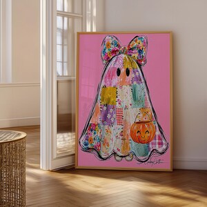 May include: A framed art print on a pink background featuring a ghost character made of various colourful floral and checkered fabric patches. The ghost wears a large matching floral bow on its head and carries a small orange jack-o'-lantern bucket.